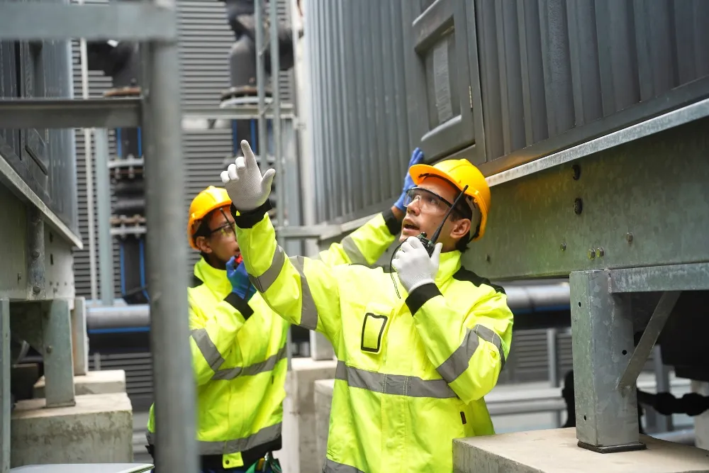 Two industrial workers wearing yellow hard hats and high-visibility jackets inspect large machinery.